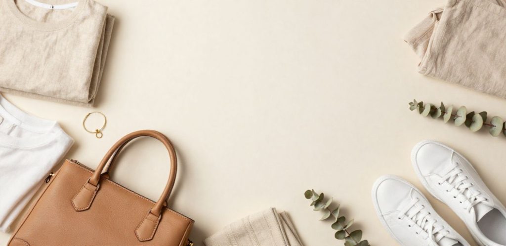 Flat lay of neutral fashion essentials including a tan leather bag, white trainers, folded beige clothing and eucalyptus sprigs on a cream background