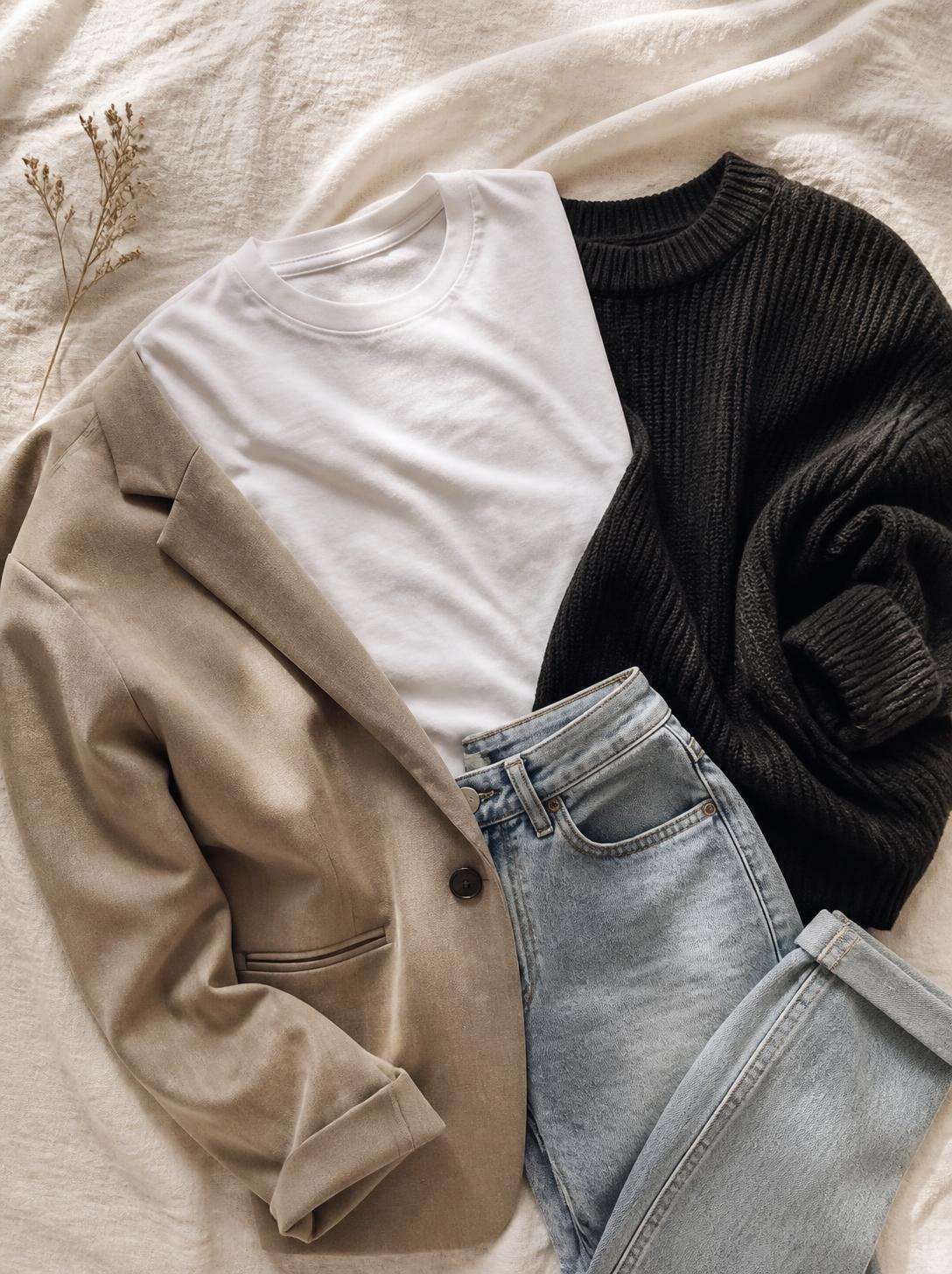 Flatlay of a white t-shirt, a black knit, jeans and a blazer on a warm cream linen surface